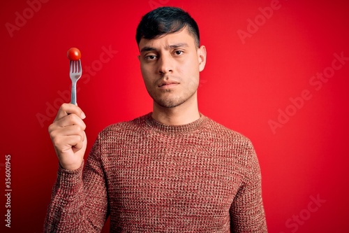 Young hispanic man holding fresh organic tomato on fork over red isolated background with a confident expression on smart face thinking serious