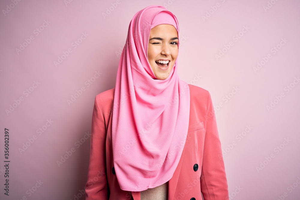 Fototapeta premium Young beautiful girl wearing muslim hijab standing over isolated pink background winking looking at the camera with sexy expression, cheerful and happy face.