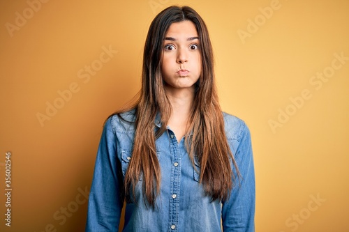 Young beautiful girl wearing casual denim shirt standing over isolated yellow background puffing cheeks with funny face. Mouth inflated with air, crazy expression. © Krakenimages.com