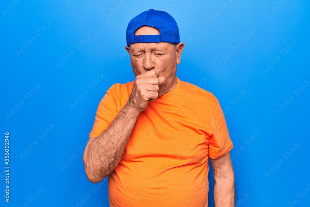 Senior handsome grey-haired man wearing casual t-shirt and cap over blue background feeling unwell and coughing as symptom for cold or bronchitis. Health care concept.