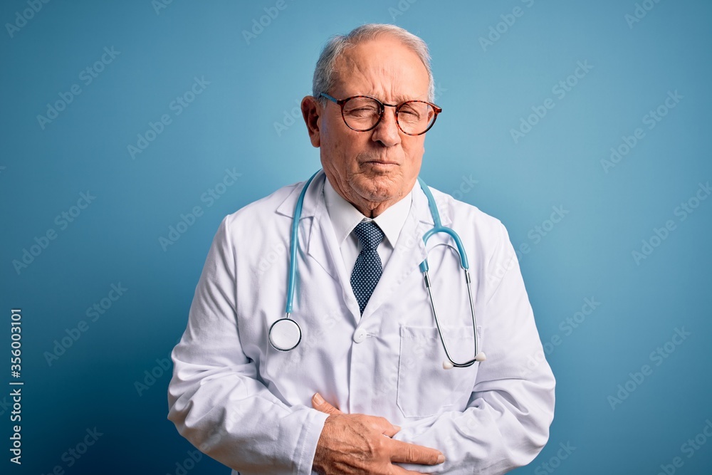 Senior grey haired doctor man wearing stethoscope and medical coat over blue background with hand on stomach because indigestion, painful illness feeling unwell. Ache concept. Senior grey haired doctor man wearing stethoscope and medical coat over blue background with hand on stomach because indigestion, painful illness feeling unwell. Ache concept.