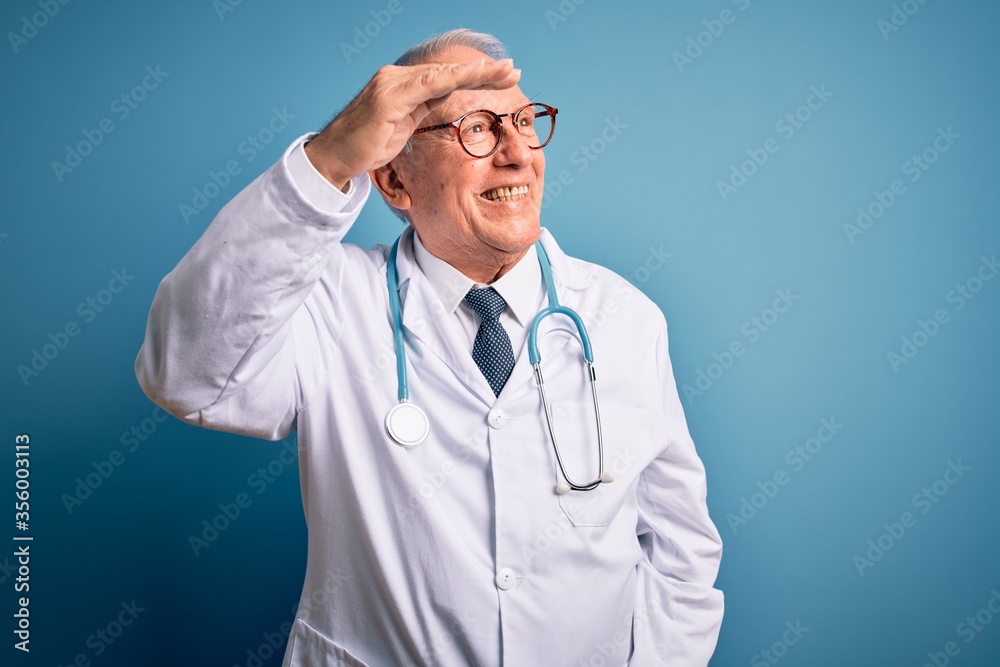 Senior grey haired doctor man wearing stethoscope and medical coat over blue background very happy and smiling looking far away with hand over head. Searching concept. Senior grey haired doctor man wearing stethoscope and medical coat over blue background very happy and smiling looking far away with hand over head. Searching concept.