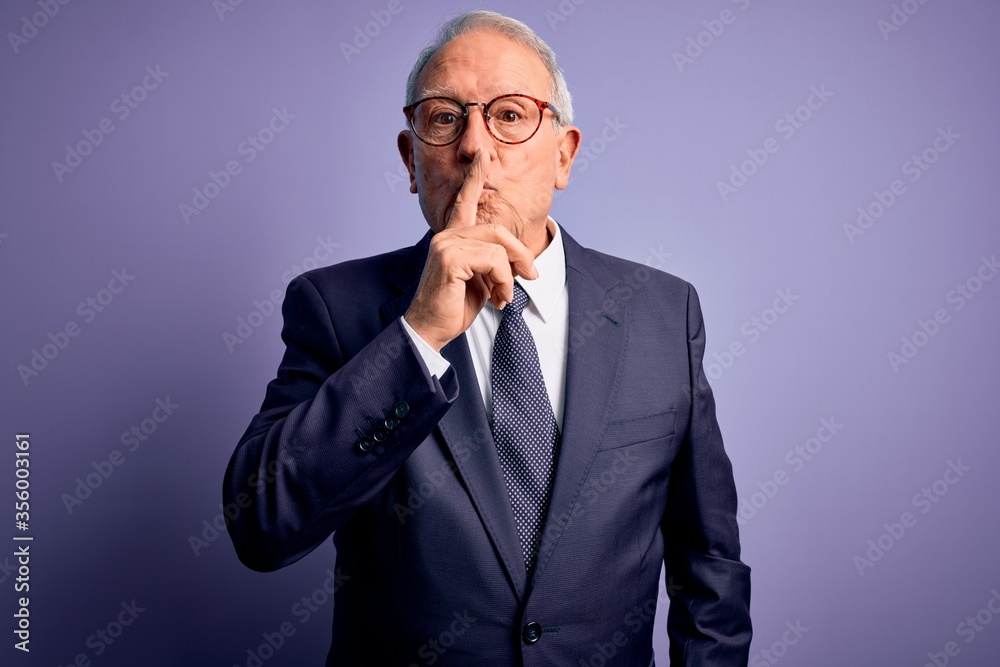 Grey haired senior business man wearing glasses and elegant suit and tie over purple background asking to be quiet with finger on lips. Silence and secret concept. Grey haired senior business man wearing glasses and elegant suit and tie over purple background asking to be quiet with finger on lips. Silence and secret concept.