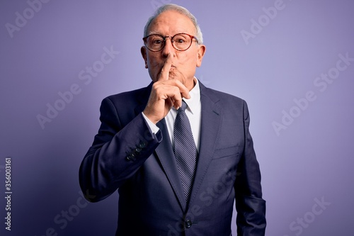 Grey haired senior business man wearing glasses and elegant suit and tie over purple background asking to be quiet with finger on lips. Silence and secret concept.