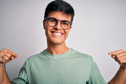 Young handsome man wearing casual t-shirt and glasses over isolated white background looking confident with smile on face, pointing oneself with fingers proud and happy.