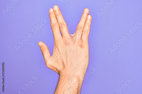 Hand of caucasian young man showing fingers over isolated purple background greeting doing Vulcan salute, showing back of the hand and fingers, freak culture