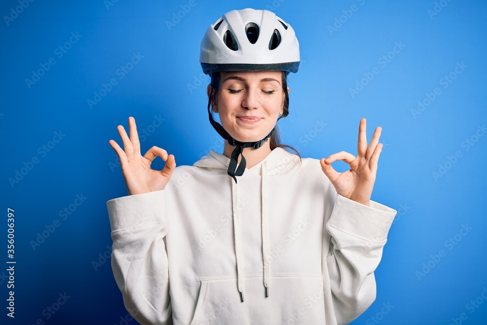 Young beautiful redhead cyclist woman wearing bike helmet over isolated blue background relax and smiling with eyes closed doing meditation gesture with fingers. Yoga concept. Young beautiful redhead cyclist woman wearing bike helmet over isolated blue background relax and smiling with eyes closed doing meditation gesture with fingers. Yoga concept.