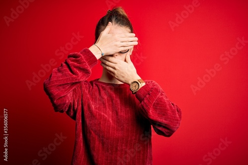 Young beautiful redhead woman wearing casual sweater over isolated red background Covering eyes and mouth with hands, surprised and shocked. Hiding emotion © Krakenimages.com