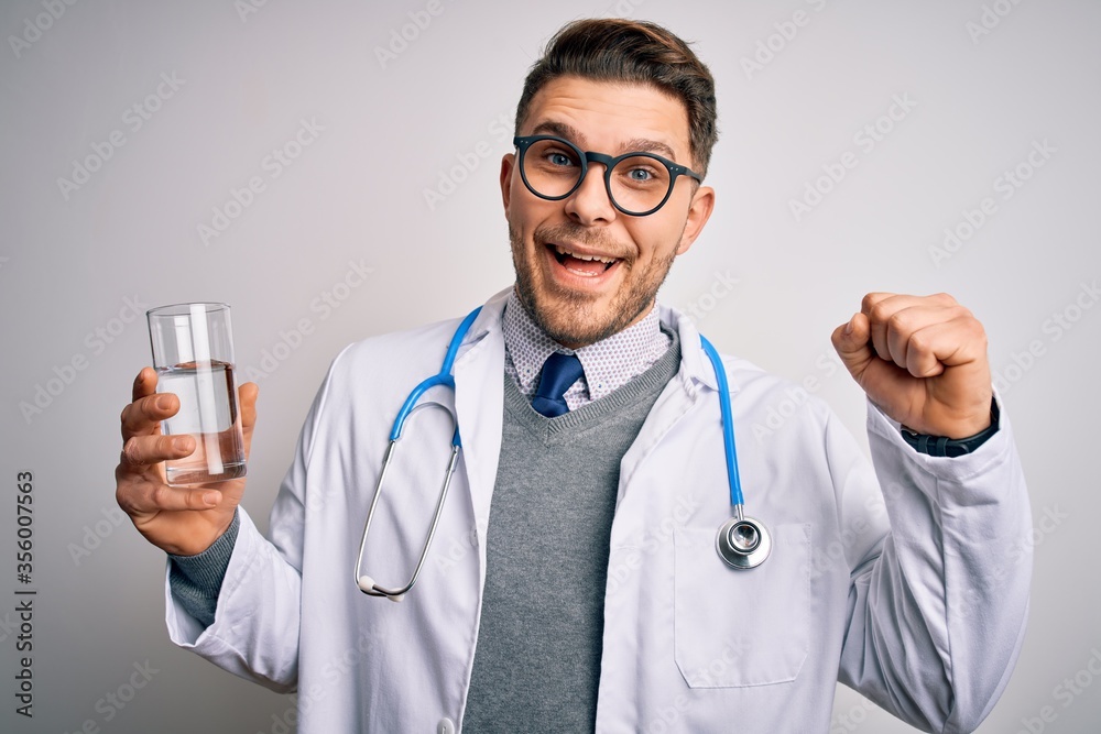 Young doctor man wearing medical coat holding a glass of fresh water over isolated background screaming proud and celebrating victory and success very excited, cheering emotion
