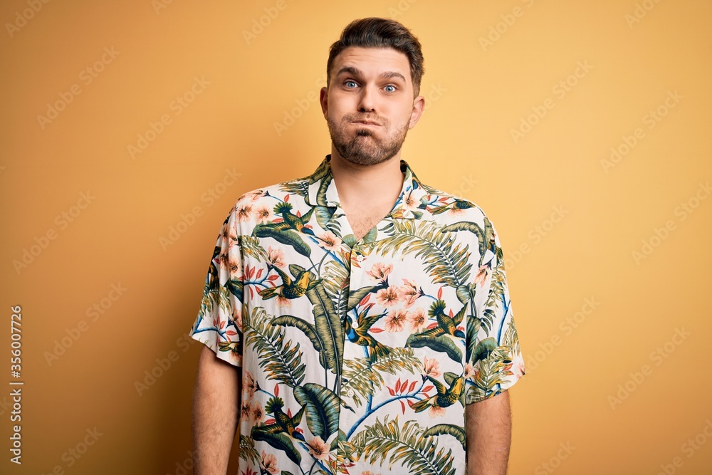 Young man with blue eyes on vacation wearing floral summer shirt over yellow background puffing cheeks with funny face. Mouth inflated with air, crazy expression. Young man with blue eyes on vacation wearing floral summer shirt over yellow background puffing cheeks with funny face. Mouth inflated with air, crazy expression.