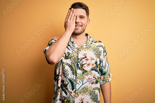 Young man with blue eyes on vacation wearing floral summer shirt over yellow background covering one eye with hand  confident smile on face and surprise emotion.