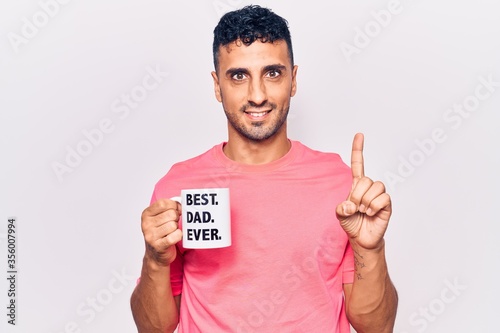 Young hispanic man drinking mug of coffe with best dad ever message smiling with an idea or question pointing finger with happy face, number one