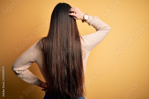 Young beautiful asian woman wearing casual sweater over yellow isolated background Backwards thinking about doubt with hand on head