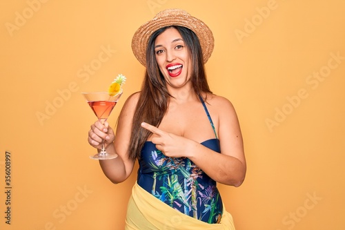Young hispanic woman wearing summer swimsuit drinking a cocktail over yellow background very happy pointing with hand and finger