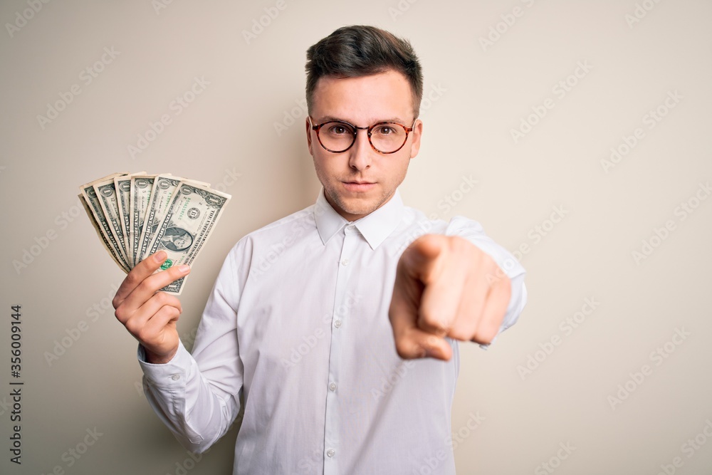 Young handsome caucasian business man holding bunch of one dollar banknotes pointing with finger to the camera and to you, hand sign, positive and confident gesture from the front Young handsome caucasian business man holding bunch of one dollar banknotes pointing with finger to the camera and to you, hand sign, positive and confident gesture from the front