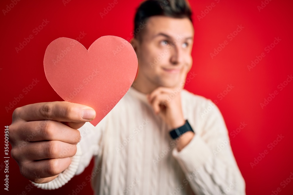 Young handsome caucasian man holding paper shape heart over red background serious face thinking about question, very confused idea Young handsome caucasian man holding paper shape heart over red background serious face thinking about question, very confused idea