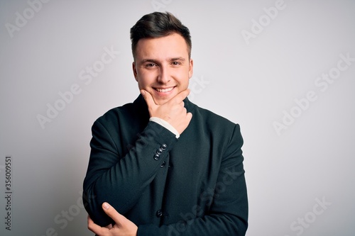 Young handsome business mas wearing elegant winter coat standing over isolated background looking confident at the camera smiling with crossed arms and hand raised on chin. Thinking positive.