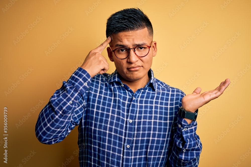 Young handsome latin man wearing casual shirt and glasses over yellow background confused and annoyed with open palm showing copy space and pointing finger to forehead. Think about it. Young handsome latin man wearing casual shirt and glasses over yellow background confused and annoyed with open palm showing copy space and pointing finger to forehead. Think about it.