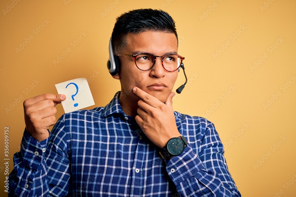 Young latin call center agent man holding question mark reminder paper using headset serious face thinking about question, very confused idea Young latin call center agent man holding question mark reminder paper using headset serious face thinking about question, very confused idea