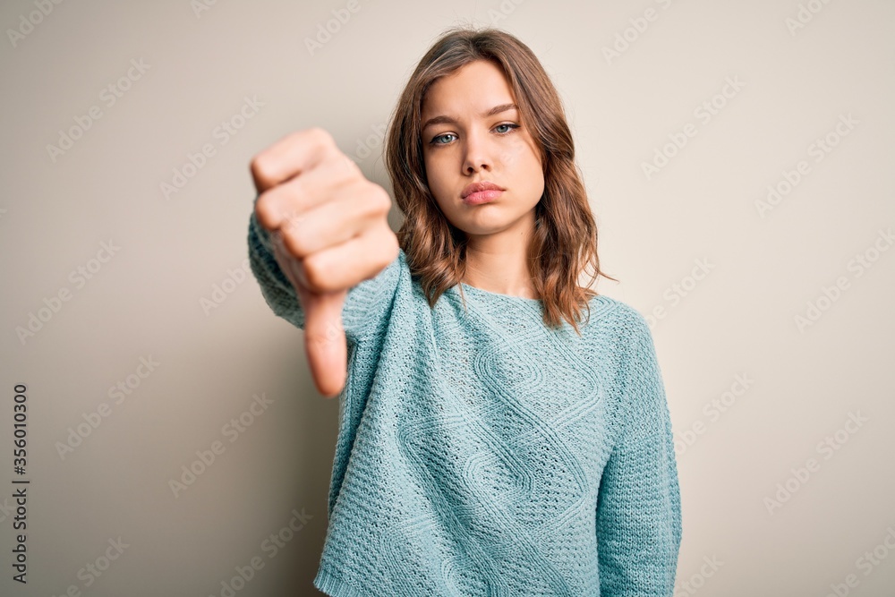 Young blonde girl wearing casual blue winter sweater over isolated background looking unhappy and angry showing rejection and negative with thumbs down gesture. Bad expression. Young blonde girl wearing casual blue winter sweater over isolated background looking unhappy and angry showing rejection and negative with thumbs down gesture. Bad expression.