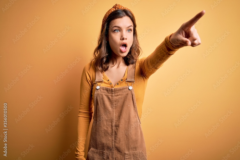 Young beautiful blonde girl wearing overall standing over yellow isolated background Pointing with finger surprised ahead, open mouth amazed expression, something on the front