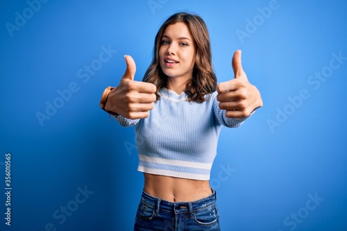 Young beautiful blonde girl wearing casual sweater standing over blue isolated background approving doing positive gesture with hand  thumbs up smiling and happy for success. Winner gesture.