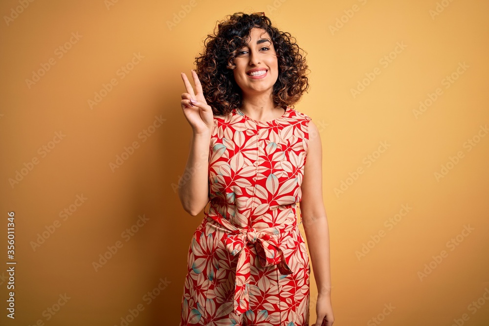 Young beautiful curly arab woman on vacation wearing summer floral dress and sunglasses showing and pointing up with fingers number two while smiling confident and happy.