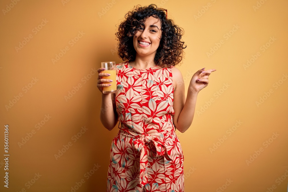 Young beautiful curly arab woman drinking glass of healthy orange juice over yellow background very happy pointing with hand and finger to the side Young beautiful curly arab woman drinking glass of healthy orange juice over yellow background very happy pointing with hand and finger to the side