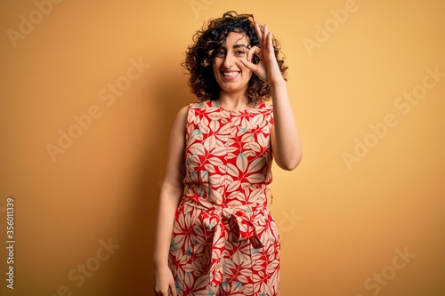 Young beautiful curly arab woman on vacation wearing summer floral dress and sunglasses smiling positive doing ok sign with hand and fingers. Successful expression.