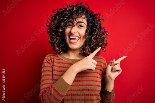Young beautiful curly arab woman wearing casual striped sweater standing over red background smiling and looking at the camera pointing with two hands and fingers to the side.