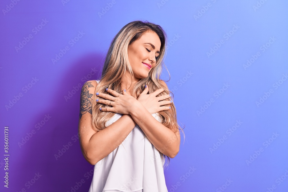 Fototapeta premium Young beautiful blonde woman wearing white t-shirt standing over isolated purple background hugging oneself happy and positive, smiling confident. Self love and self care