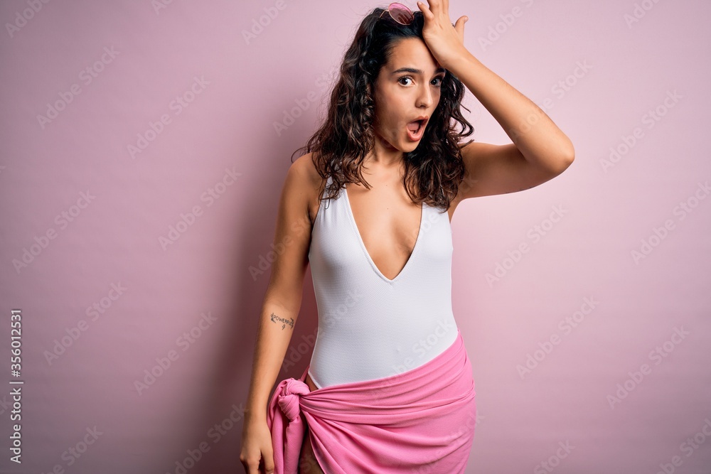 Beautiful woman with curly hair on vacation wearing white swimsuit over pink background surprised with hand on head for mistake, remember error. Forgot, bad memory concept. Beautiful woman with curly hair on vacation wearing white swimsuit over pink background surprised with hand on head for mistake, remember error. Forgot, bad memory concept.