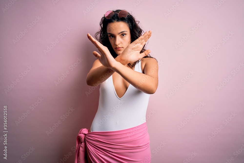 Beautiful woman with curly hair on vacation wearing white swimsuit over pink background Rejection expression crossing arms doing negative sign, angry face Beautiful woman with curly hair on vacation wearing white swimsuit over pink background Rejection expression crossing arms doing negative sign, angry face
