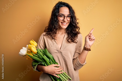 Young beautiful romantic woman with curly hair holding bouquet of yellow tulips cheerful with a smile on face pointing with hand and finger up to the side with happy and natural expression
