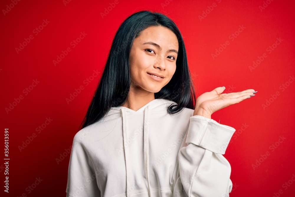 Young beautiful chinese sporty woman wearing sweatshirt over isolated red background smiling cheerful presenting and pointing with palm of hand looking at the camera. Young beautiful chinese sporty woman wearing sweatshirt over isolated red background smiling cheerful presenting and pointing with palm of hand looking at the camera.