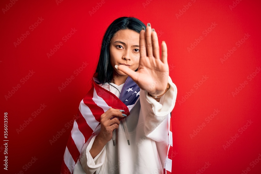 Obraz premium Young beautiful chinese woman wearing United States flag over isolated red background with open hand doing stop sign with serious and confident expression, defense gesture