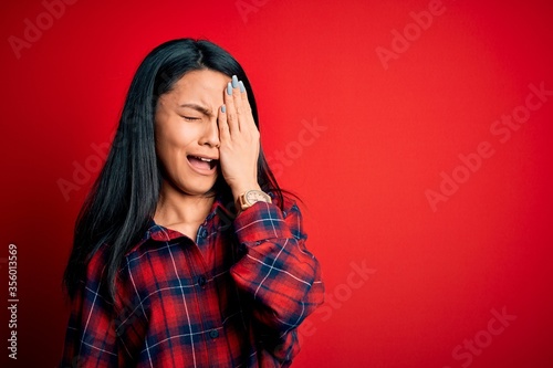 Young beautiful chinese woman wearing casual shirt over isolated red background Yawning tired covering half face, eye and mouth with hand. Face hurts in pain. © Krakenimages.com
