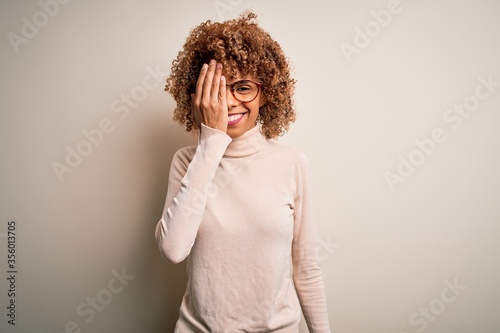 Young african american woman wearing turtleneck sweater and glasses over white background covering one eye with hand, confident smile on face and surprise emotion.