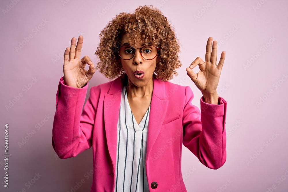 Young african american businesswoman wearing glasses standing over pink background looking surprised and shocked doing ok approval symbol with fingers. Crazy expression Young african american businesswoman wearing glasses standing over pink background looking surprised and shocked doing ok approval symbol with fingers. Crazy expression