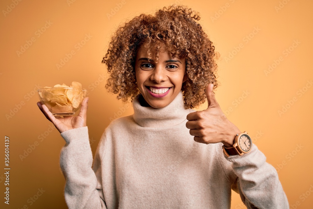 Young african american curly woman holding bowl with chips potatoes over yellow background happy with big smile doing ok sign, thumb up with fingers, excellent sign Young african american curly woman holding bowl with chips potatoes over yellow background happy with big smile doing ok sign, thumb up with fingers, excellent sign