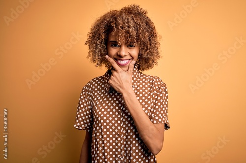 Young beautiful african american woman wearing casual shirt standing over yellow background looking confident at the camera smiling with crossed arms and hand raised on chin. Thinking positive.