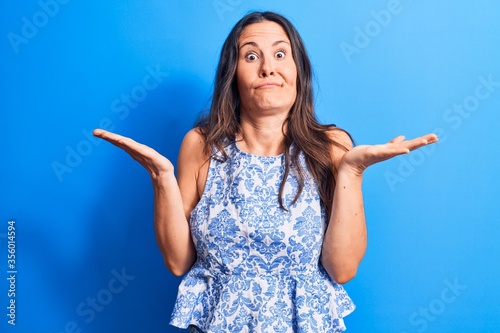 Young beautiful brunette woman wearing casual sleeveless t-shirt over blue background clueless and confused with open arms, no idea and doubtful face.