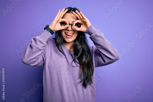 Young brunette woman wearing glasses over purple isolated background doing ok gesture like binoculars sticking tongue out, eyes looking through fingers. Crazy expression. © Krakenimages.com