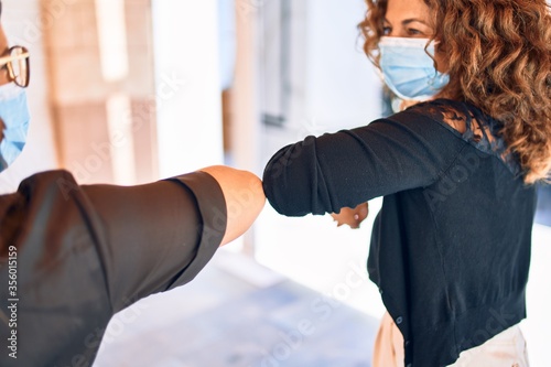 Two beautiful friends doing new elbow greeting to coronavirus prevention. Wearing medical mask standing at town street. photo