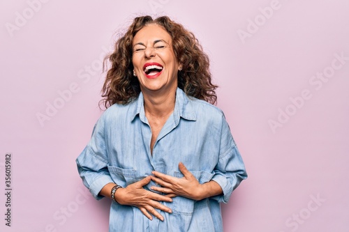 Middle age beautiful woman wearing casual denim shirt standing over pink background smiling and laughing hard out loud because funny crazy joke with hands on body Fototapet