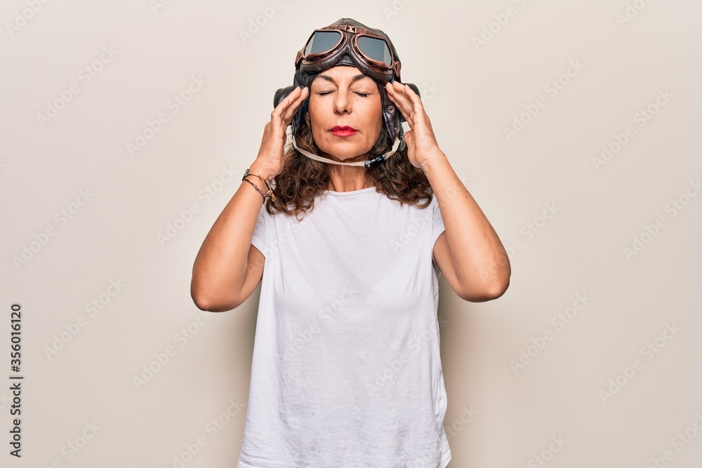 Middle age brunette woman wearing goggles and retro aviator leather hat over white background with hand on head, headache because stress. Suffering migraine. Middle age brunette woman wearing goggles and retro aviator leather hat over white background with hand on head, headache because stress. Suffering migraine.