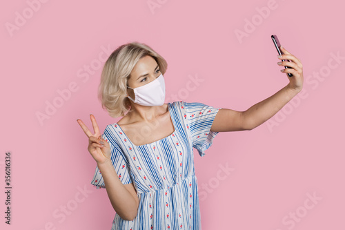 Blonde caucasian woman making a selfie gesturing the peace sign wearing a medical mask on face on a pink studio wall