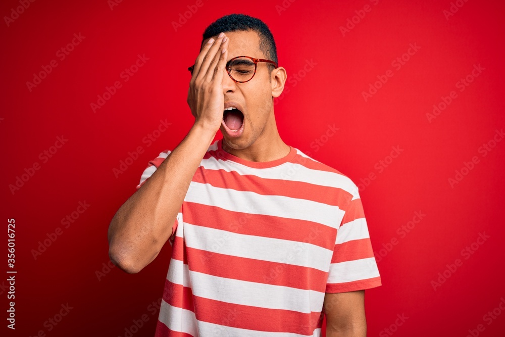 Young handsome african american man wearing casual striped t-shirt and glasses Yawning tired covering half face, eye and mouth with hand. Face hurts in pain. Young handsome african american man wearing casual striped t-shirt and glasses Yawning tired covering half face, eye and mouth with hand. Face hurts in pain.