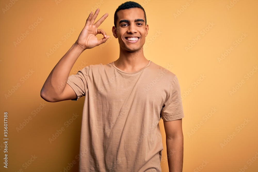 Young handsome african american man wearing casual t-shirt standing over yellow background smiling positive doing ok sign with hand and fingers. Successful expression. Young handsome african american man wearing casual t-shirt standing over yellow background smiling positive doing ok sign with hand and fingers. Successful expression.