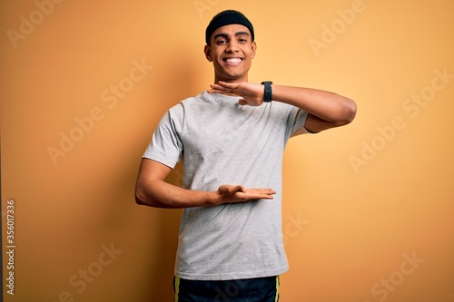 Handsome african american sportsman doing sport wearing sportswear over yellow background gesturing with hands showing big and large size sign, measure symbol. Smiling looking at the camera. Measuring